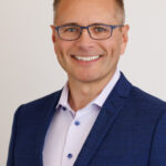 Smiling man in blue suit jacket and light dress shirt, wearing blue rectangular glasses, posed against a light neutral background.