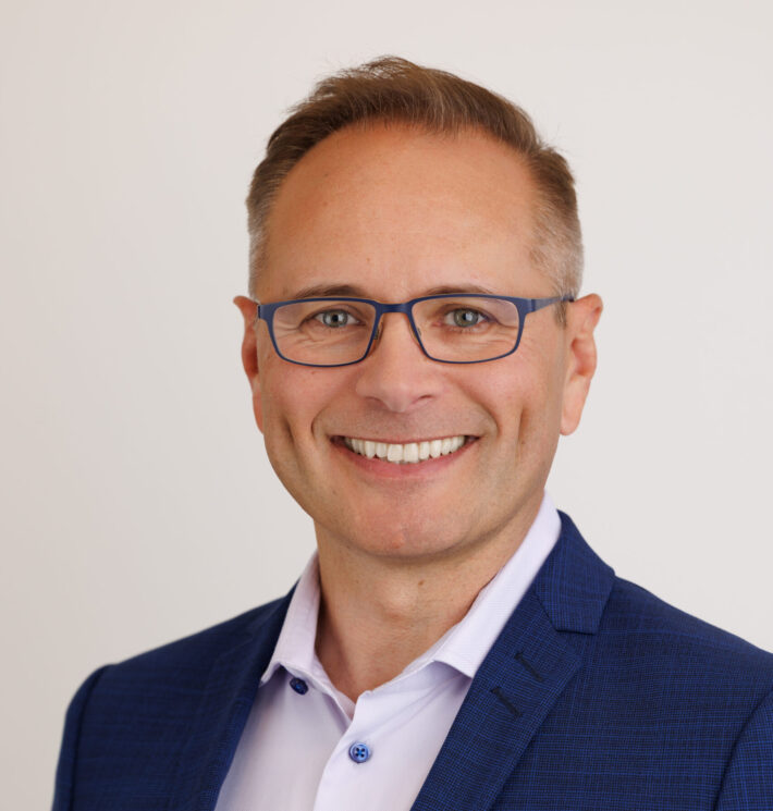 Smiling man in blue suit jacket and light dress shirt, wearing blue rectangular glasses, posed against a light neutral background.